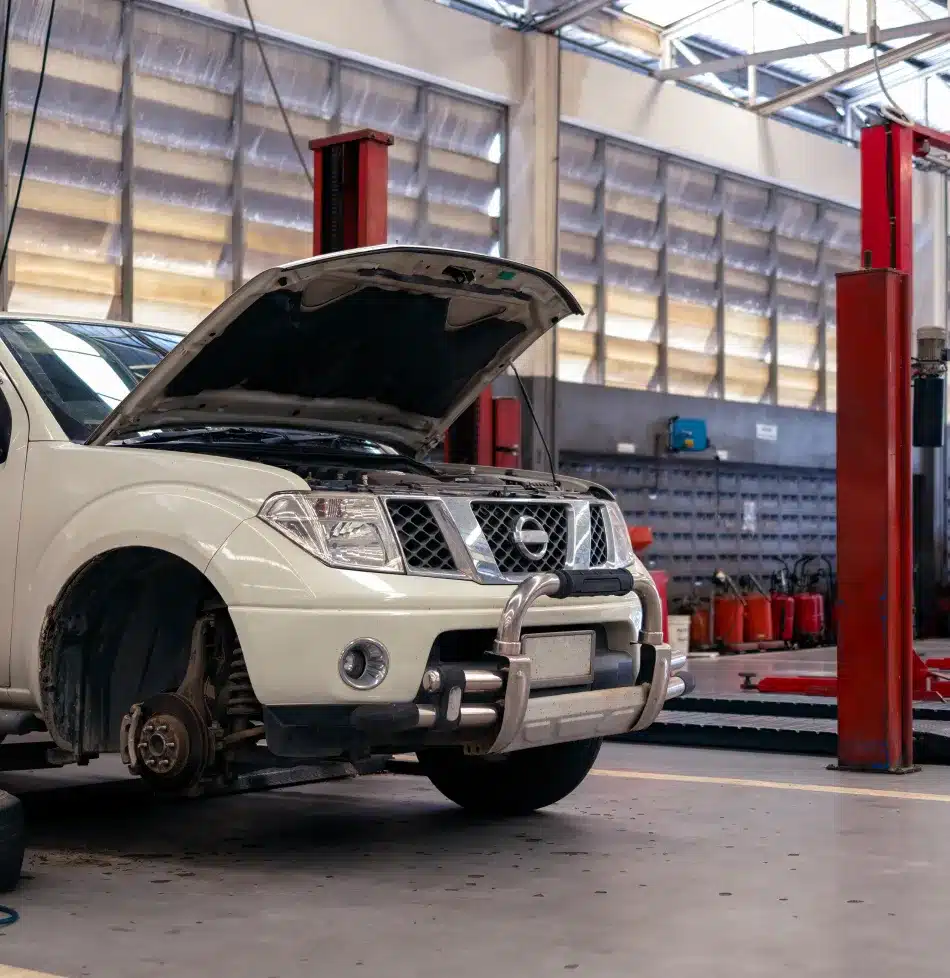 A white pickup truck with its hood open and front wheel removed sits in an auto repair shop, surrounded by equipment and machinery.