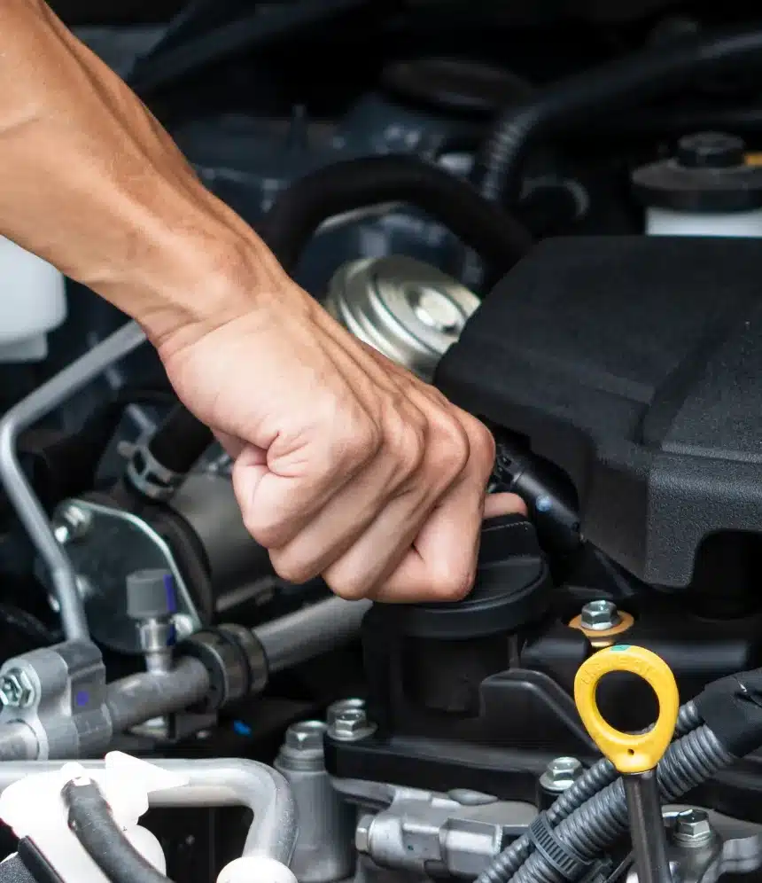 A persons hand is turning the oil cap to open it on a car engine, preparing for vehicle maintenance or an oil change. Various engine components and hoses are visible.