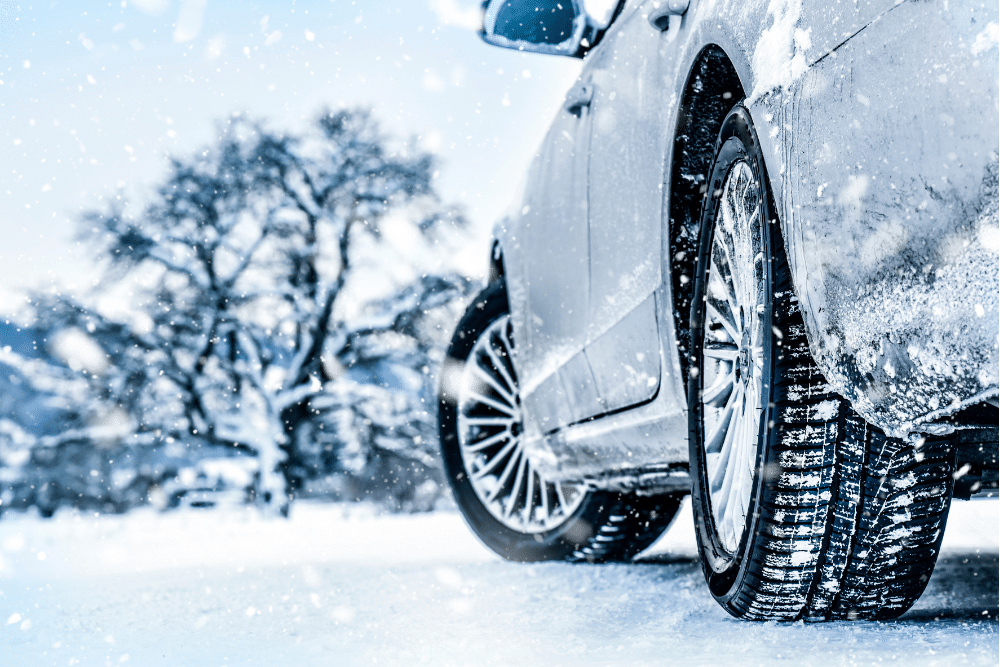 Tire Services by Pristine Motorsports in 950 S Dupont Blvd, Smyrna, DE. Close-up of a car driving on a snowy road with snow-covered tires and trees in the background.