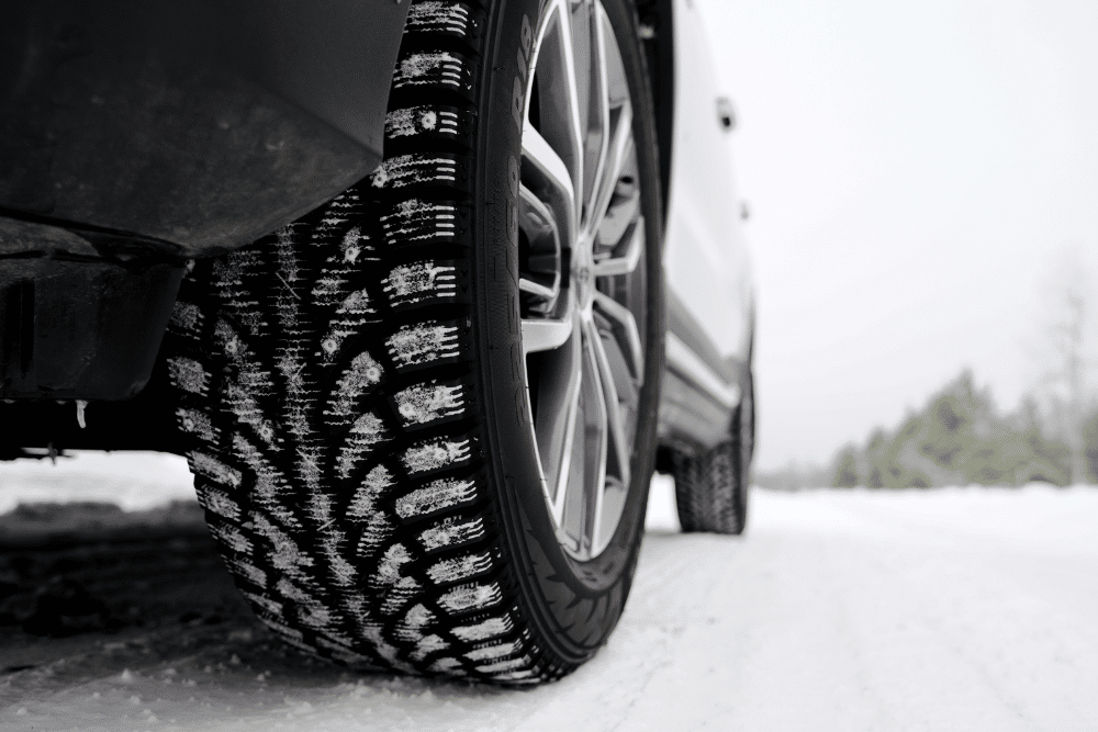 Winter vs. All-Season Tires by Pristine Motorsports in 950 S Dupont Blvd, Smyrna, DE. Close-up of a winter tire on a car driving through snow, showcasing the tread pattern for traction in snowy conditions.